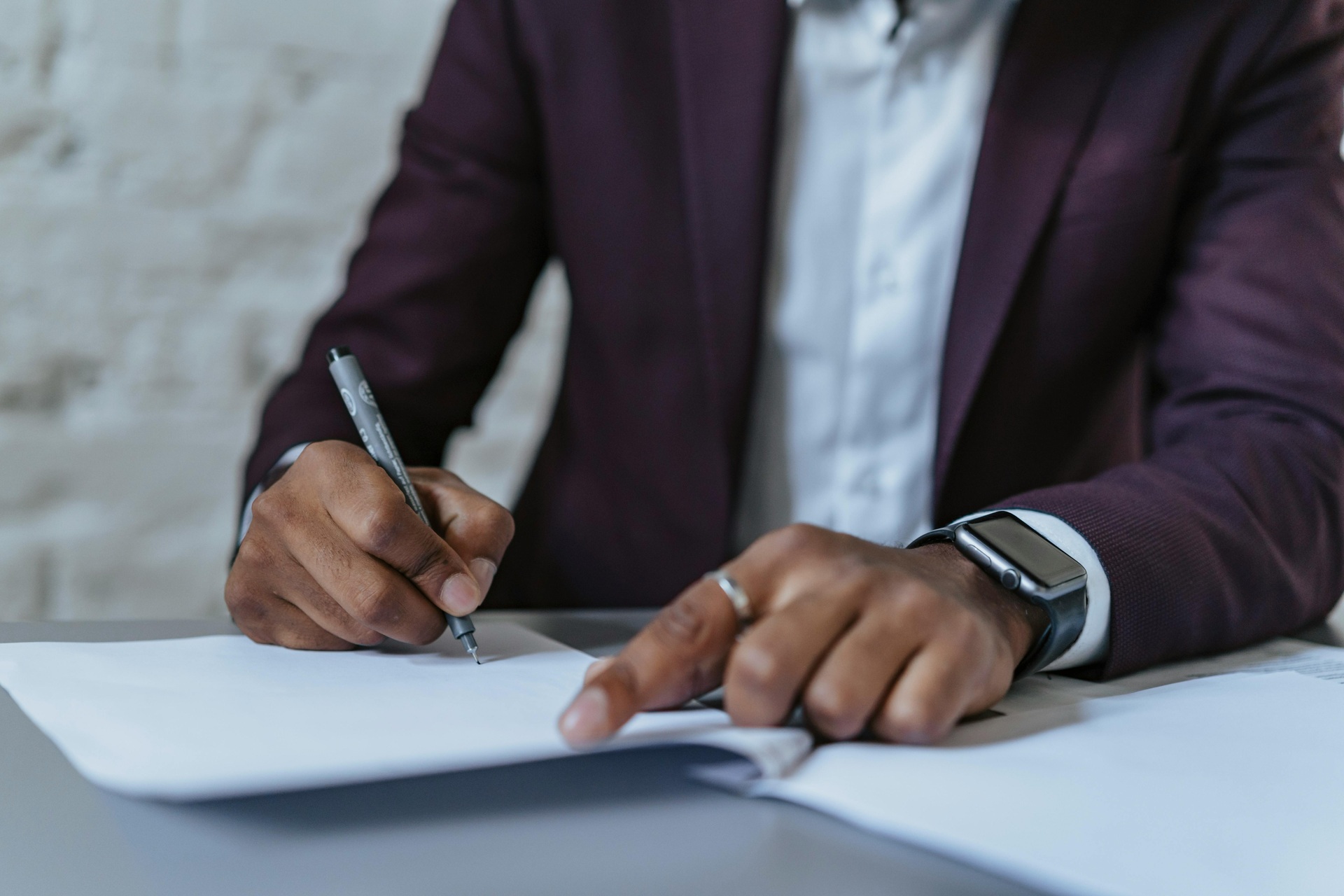 Close-up of a person in a burgundy suit jacket and white shirt signing a document with a gray pen. Close-up of a person in a burgundy suit jacket and white shirt signing a document with a gray pen.