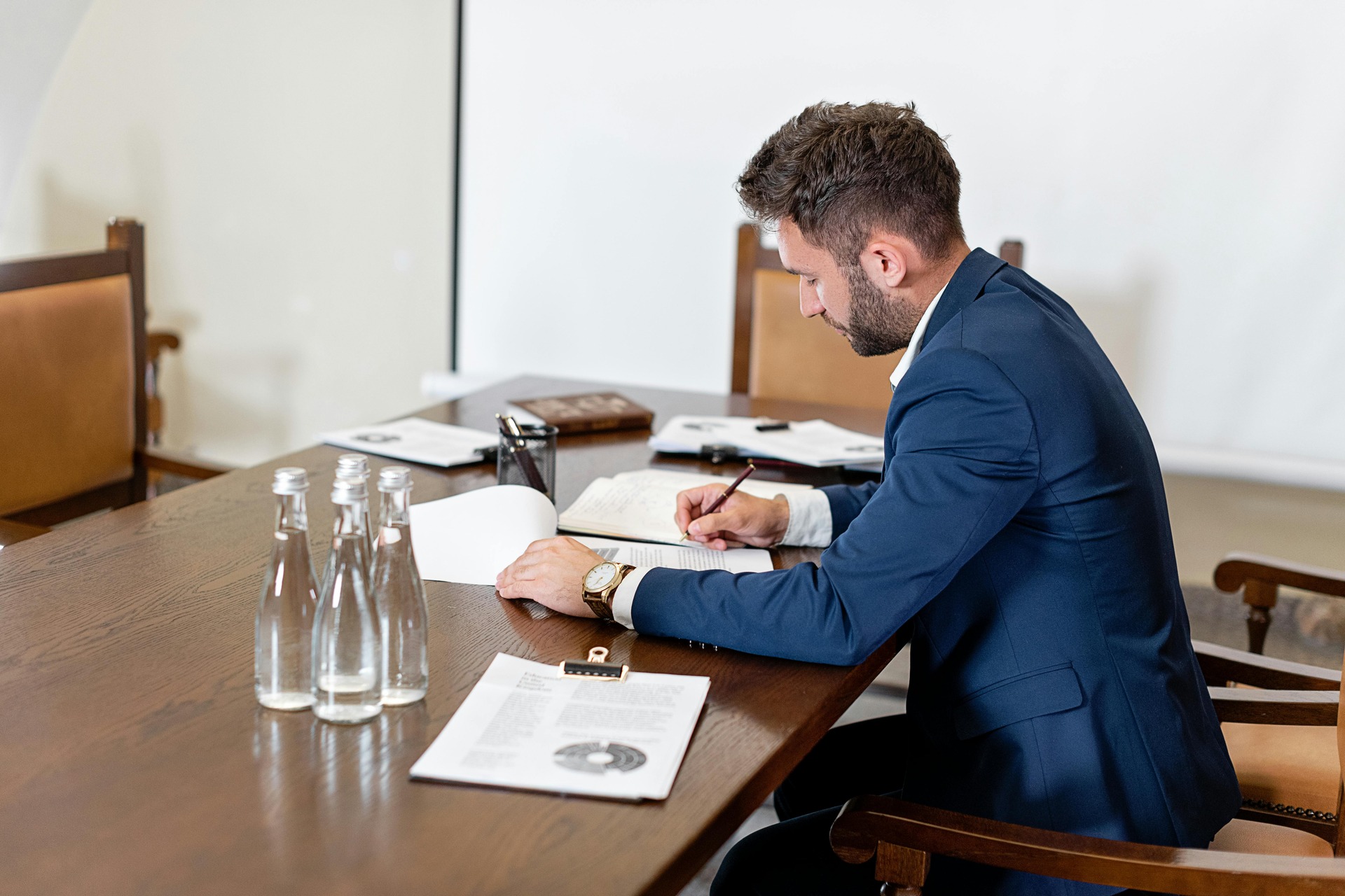 Man in a navy blue suit seated at a conference table taking notes in an open notebook. Man in a navy blue suit seated at a conference table taking notes in an open notebook.
