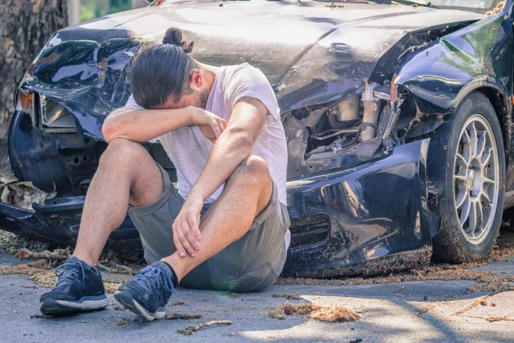 Man sitting on the ground in distress after a car accident with a heavily damaged vehicle