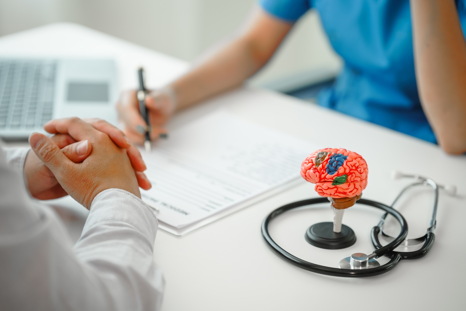 A doctor in a white coat sitting at a white table with a patient whose hands are clasped. Another person, possibly a medical professional, wearing blue scrubs, is writing on a form. A model of a human brain on a stand and a stethoscope are on the table