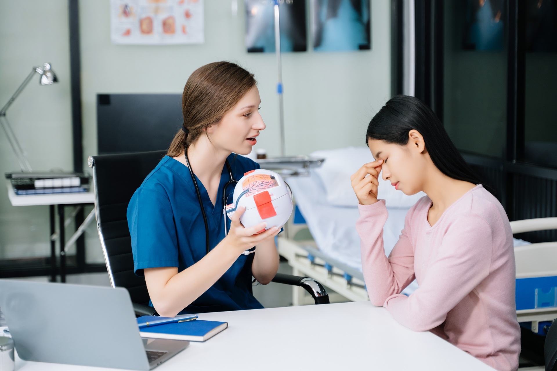 A fair-skinned female medical professional in blue scrubs holding a model of a human eye and talking to a dark-haired female patient in a pink sweater who is touching her forehead